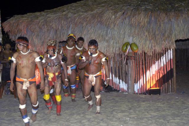 índios com microfones em frente a uma oca